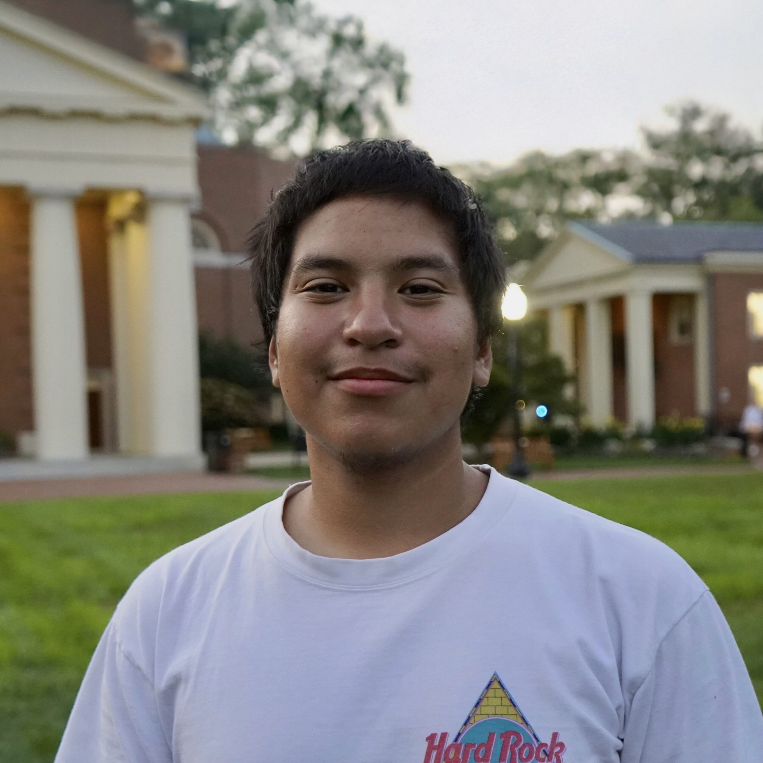 Closeup of ADC Intern David Bahena-Gutierrez smiling with green grass and brick buildings with columns in the background.